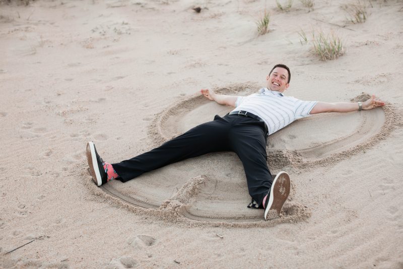 John Hawley grinning while making a sand angel in slacks, polo, and pink flamingo socks